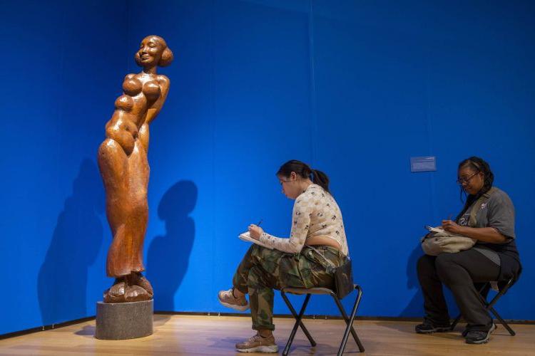photo of two women sitting in art gallery sketching on small pads of paper. A tall wood figurative sculpture is on the right side of the frame.