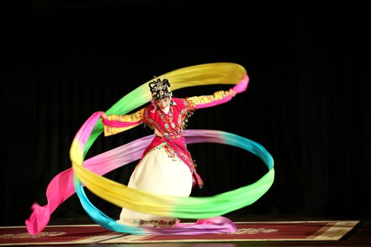 A single dancer in a white embroidered dress and red embroidered jacket, with detailed face painting and headress, dancing on a stage, twirling long colorful ribbons, one in each hand, swirling around her body. 